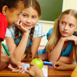 Three children in a classroom, two girls sitting and listening attentively.