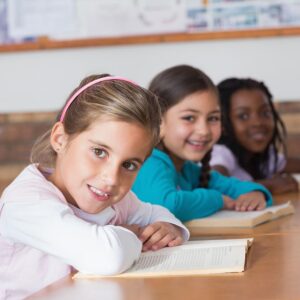 Three young girls smiling and sitting together in a classroom.