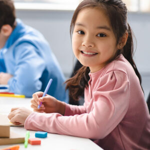 A young girl smiling at the camera while writing.