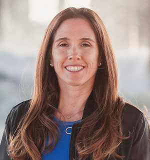 A smiling woman with long brown hair, wearing a blue top and black jacket, posing outdoors.