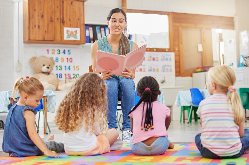 A teacher reading a book to a group of children sitting in front of her in a classroom.